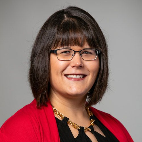 Woman with Dark brown hair, fair complexion, and black thin rim glasses, wearing a black and gold blouse with a red cardigan sweater