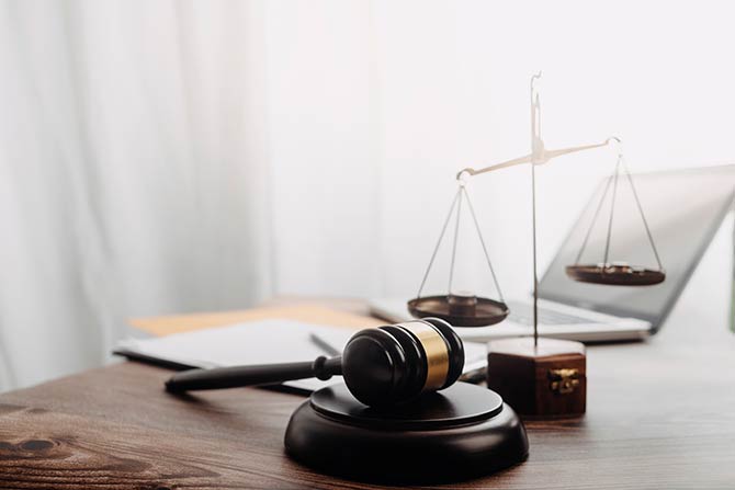 A Gavel, papers, and set of balance scales sits on a desk or table backlit by a bright window that shines on the objects.