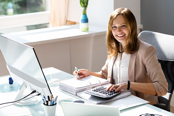 NESCPA at a Glance; A smiling woman in business attire sits at a desk with a computer, using a calculator. Sunlight streams in, creating a bright, professional atmosphere.