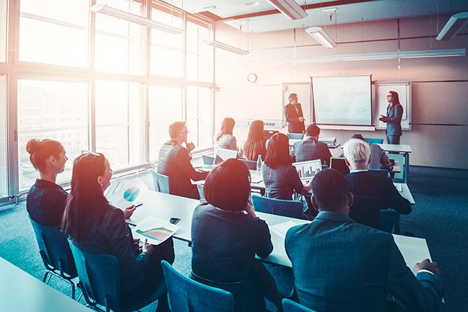 Educating Nebraska’s CPAs; A group of people seated in a conference room with a large window. Two presenters stand at the front, beside a screen. The atmosphere is attentive and professional.