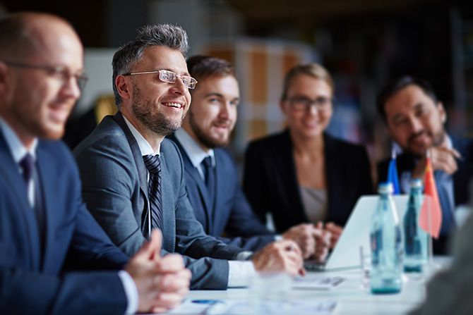 Education Your Way; A group of five people in business attire sit at a table engaged in a meeting. They appear focused and content, with water bottles and flags on the table.