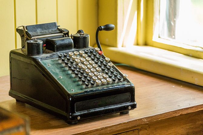 From the Beginning; A vintage mechanical calculator with numerous round keys and a lever sits on a wooden desk by a sunlit window, creating a nostalgic atmosphere.