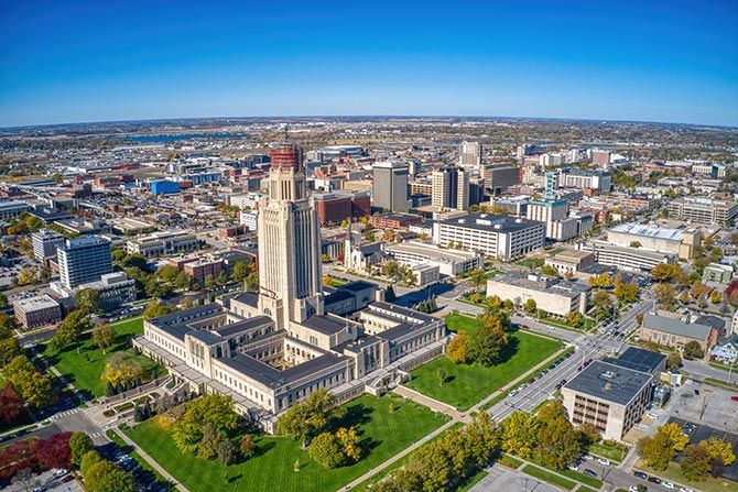 Legislative Representation Is a Priority; Aerial view of a prominent state capitol building with a tall tower surrounded by lush green lawns, set in a bustling cityscape under a clear blue sky.