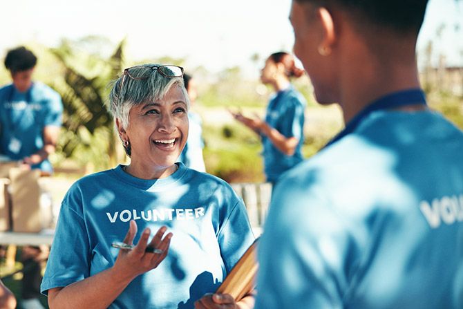 Recognizing Service to the Profession & to Nebraska; Smiling woman in a blue "Volunteer" shirt talks to another volunteer outdoors. Sunlit scene conveys teamwork and positivity.