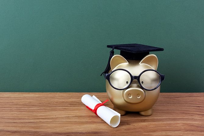 Scholarships Grow the CPA Pipeline; A gold piggy bank wearing glasses and a graduation cap sits on a wooden surface, next to a rolled diploma tied with a red ribbon, against a green backdrop.