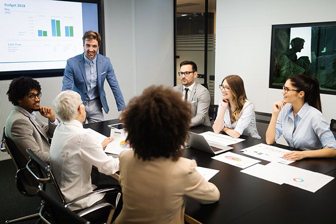 Volunteer CPAs Serve as Driving Force; A diverse group of people sit around a conference table in a modern office, engaged in a meeting. A presenter stands, smiling, near a projected graph.