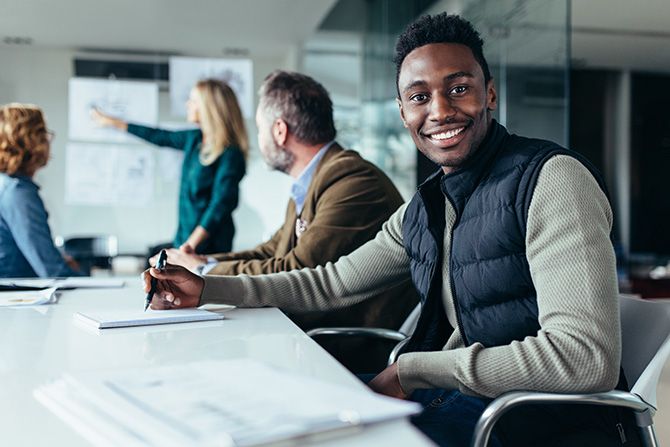 Volunteer to Make a Difference; Man smiling at a conference table, pen in hand. Colleagues in background discuss charts, creating a collaborative and positive atmosphere.