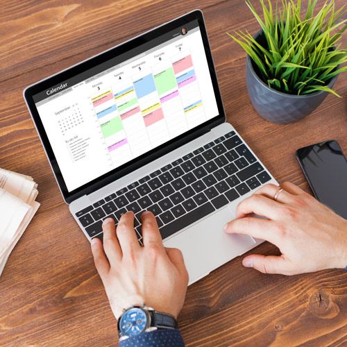 2026 NESCPA Course Calendar; A person types on a laptop displaying a colorful calendar. Nearby, a potted plant, smartphone, and stacked papers rest on a wooden desk, creating a productive workspace.