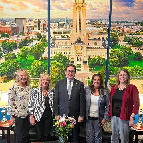 Outgoing President's Message: A Final Reflection: Strength in Membership, Strength in Purpose; Group of five people smiling in an office, with a large cityscape mural of a building behind them. Flowers are on a table in the foreground.