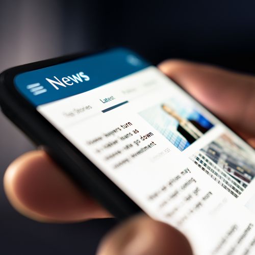 Firms in the News; Close-up of a hand holding a smartphone displaying a news app. The screen shows headlines and images, suggesting an engaged and informative tone.
