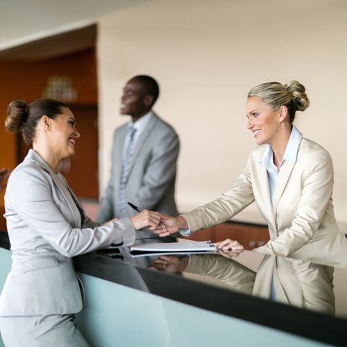 Private Banking: What Is It and Who Is It For? Two businesswomen in suits shake hands over a reception desk, smiling warmly. A businessman in the background observes, creating a professional, friendly atmosphere.