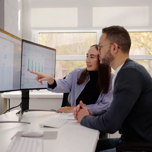 Registered Apprenticeship; Two colleagues in a well-lit office discuss data on dual monitors. The woman points at a bar graph, indicating analysis. Both appear focused and engaged.