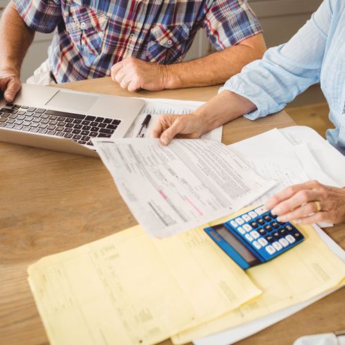Retired (or Soon Will Be)? How to Appeal Higher Medicare Premiums; Two people sitting at a table review financial documents and use a calculator and laptop, focused on paperwork. The mood is serious and focused.