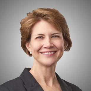 Roberta Christensen; A person with short brown hair smiles warmly at the camera, wearing a dark blazer. The background is a neutral gray, conveying a professional tone.