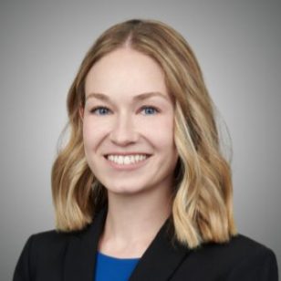 Kayla Helgoth; A smiling woman with shoulder-length blonde hair wearing a black blazer and blue top, posing against a gray background, conveys professionalism and warmth.